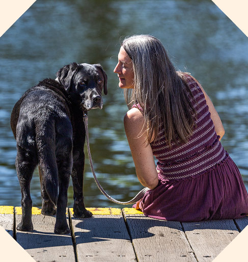 A woman sits on a wooden dock by a lake, holding a lead attached to a black dog who is standing beside her.