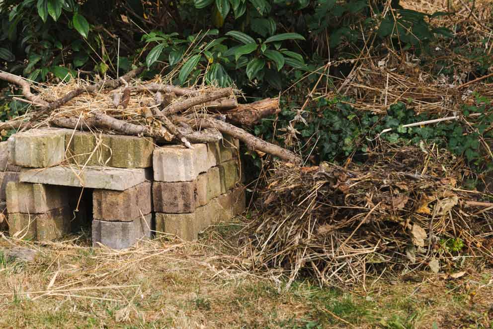 A simple hedgehog house made from stacked bricks in a grassy garden corner. The shelter has a small entrance tunnel and is topped with dry twigs and straw for insulation, blending into the natural surroundings.