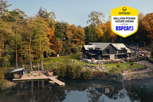 A large detached house surrounded by autumnal trees, next to a lake. A wooden hot tub and jetty sit a few metres down from the house.