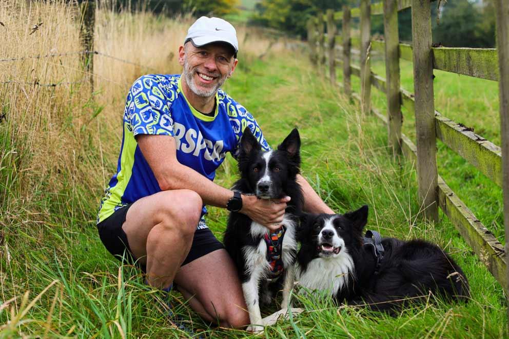 Man wearing an RSPCA branded T-shirt kneeling with two dogs in the countryside.