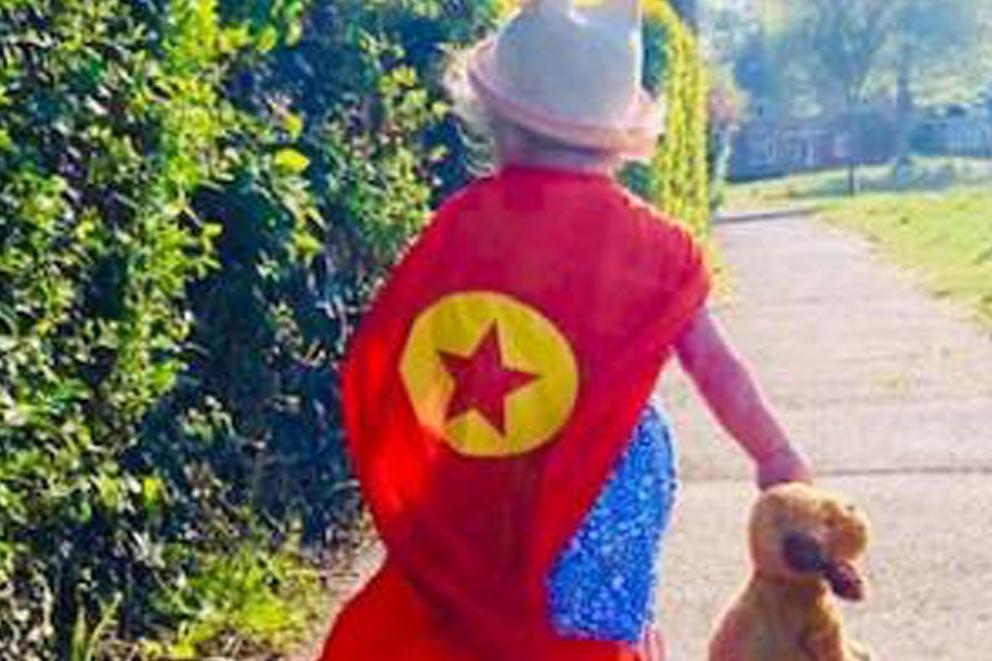 Child wearing a star patterned cape and holding a teddy bear while walking up the street.