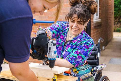 Woman in wheelchair drilling into wood, a second woman is assisting, they are both smiling